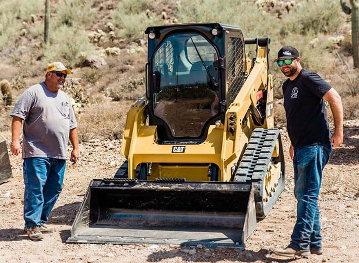 two men on dirt lot with hats standing next to a  compact track loader